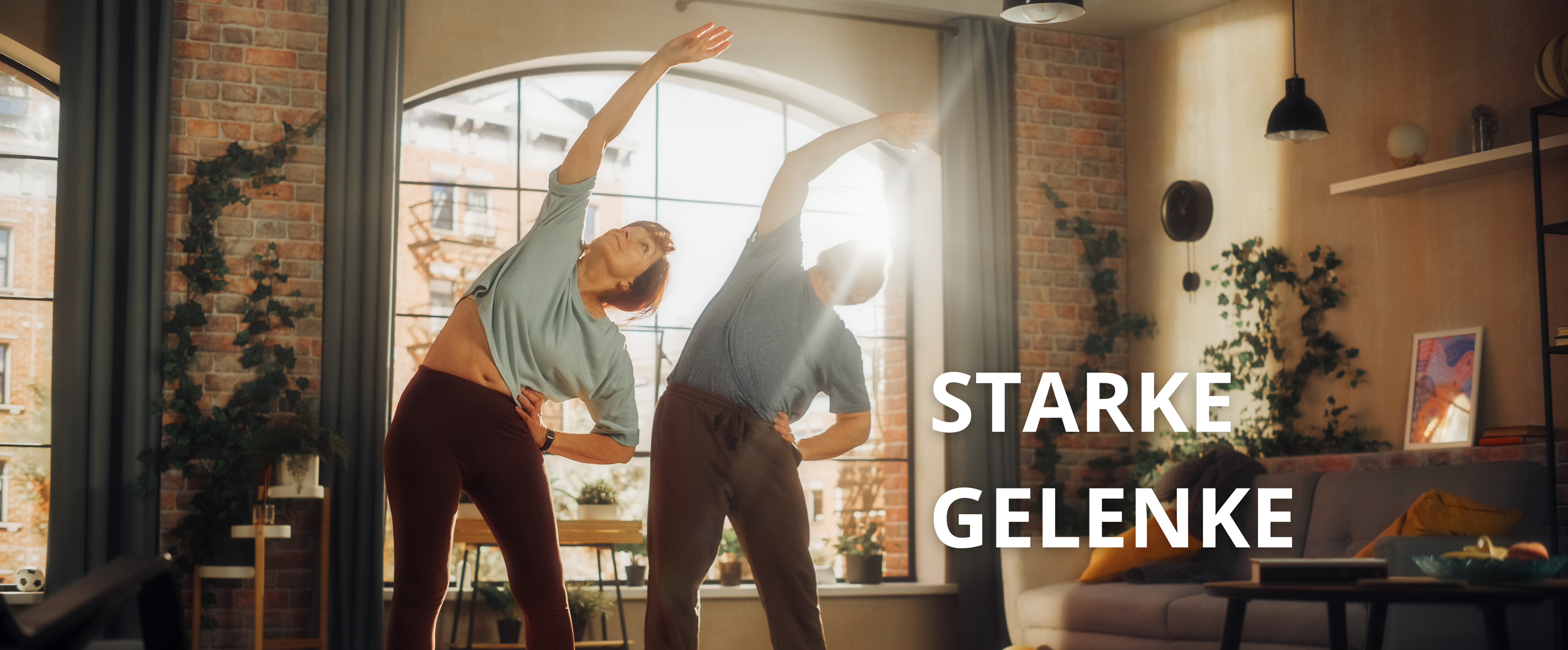 Ein Mann (rechts) und eine Frau (links) dehnen sich wobei sie den rechten Arm nach oben Strecken und dabei auf einer Yoga-Matte stehen.