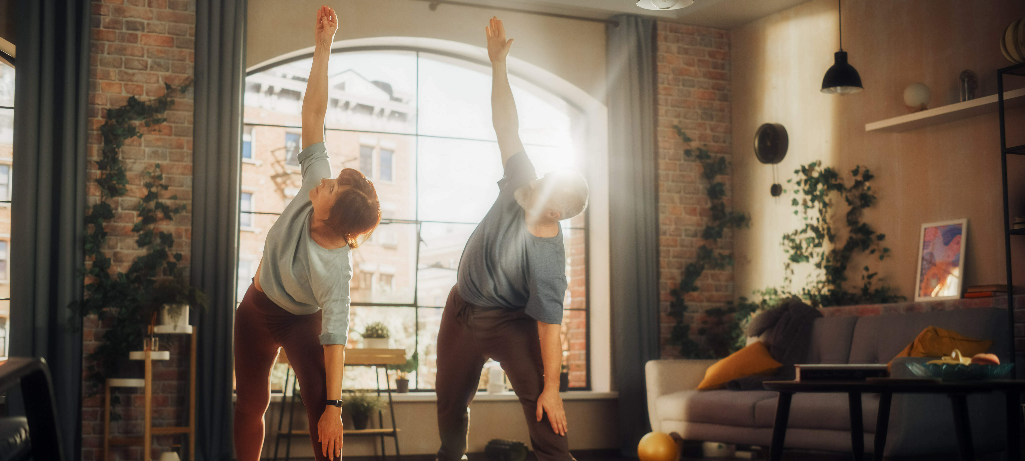 Eine Frau (links) und ein Mann (rechts) machen Yogaübungen im Wohnzimmer und heben dabei den linken Arm. Der rechte Arm liegt auf dem Oberschenkel.