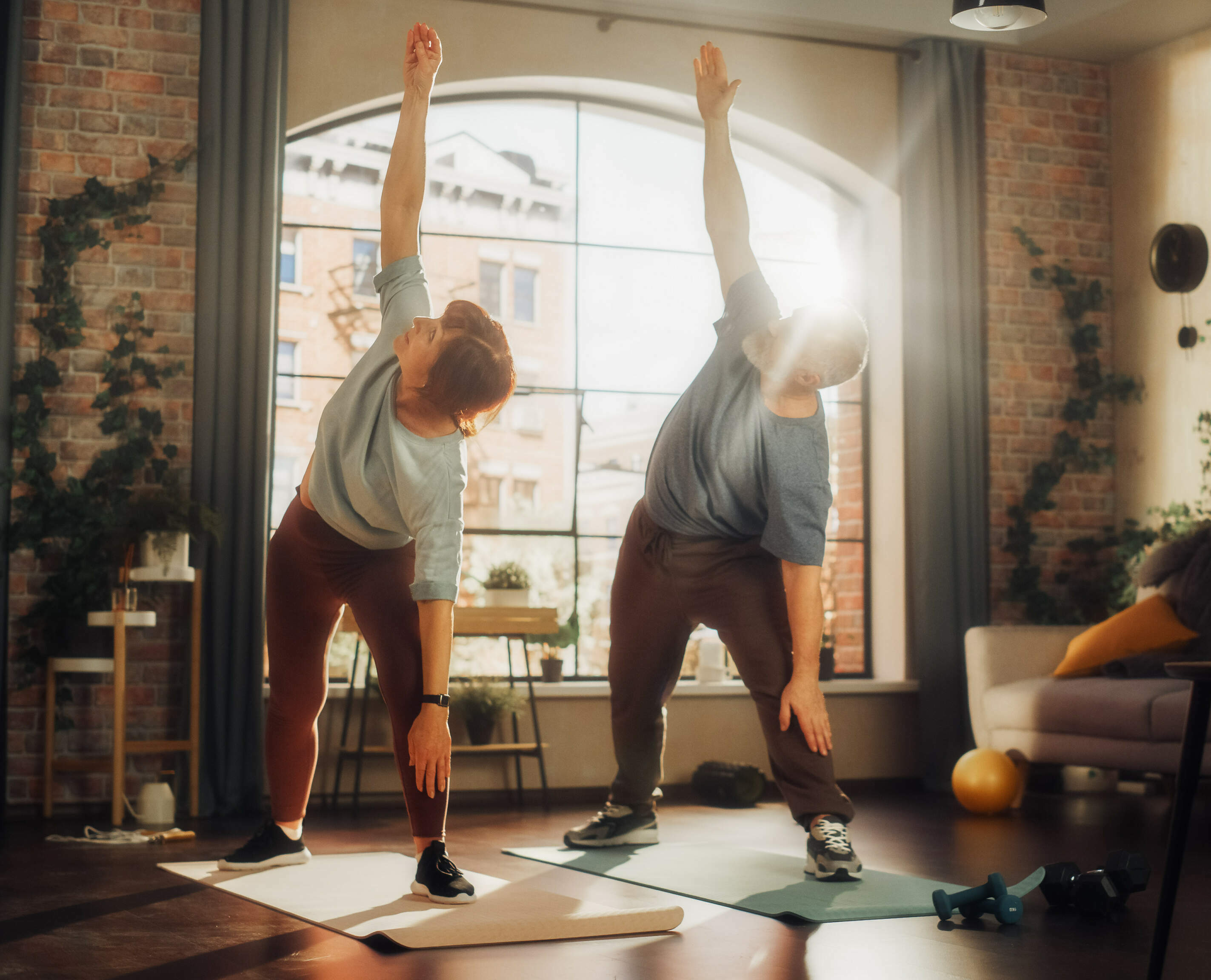 Eine Frau (links) und ein Mann (rechts) machen Yogaübungen im Wohnzimmer und heben dabei den linken Arm. Der rechte Arm liegt auf dem Oberschenkel.