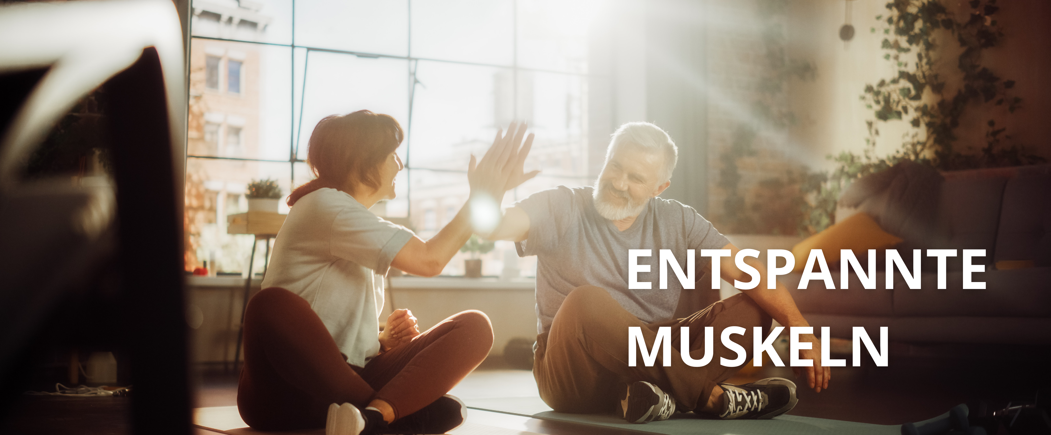 Eine Frau (links) und ein Mann (rechts) machen Yogaübungen im Wohnzimmer und heben dabei den rechten Arm um abzuklatschen.